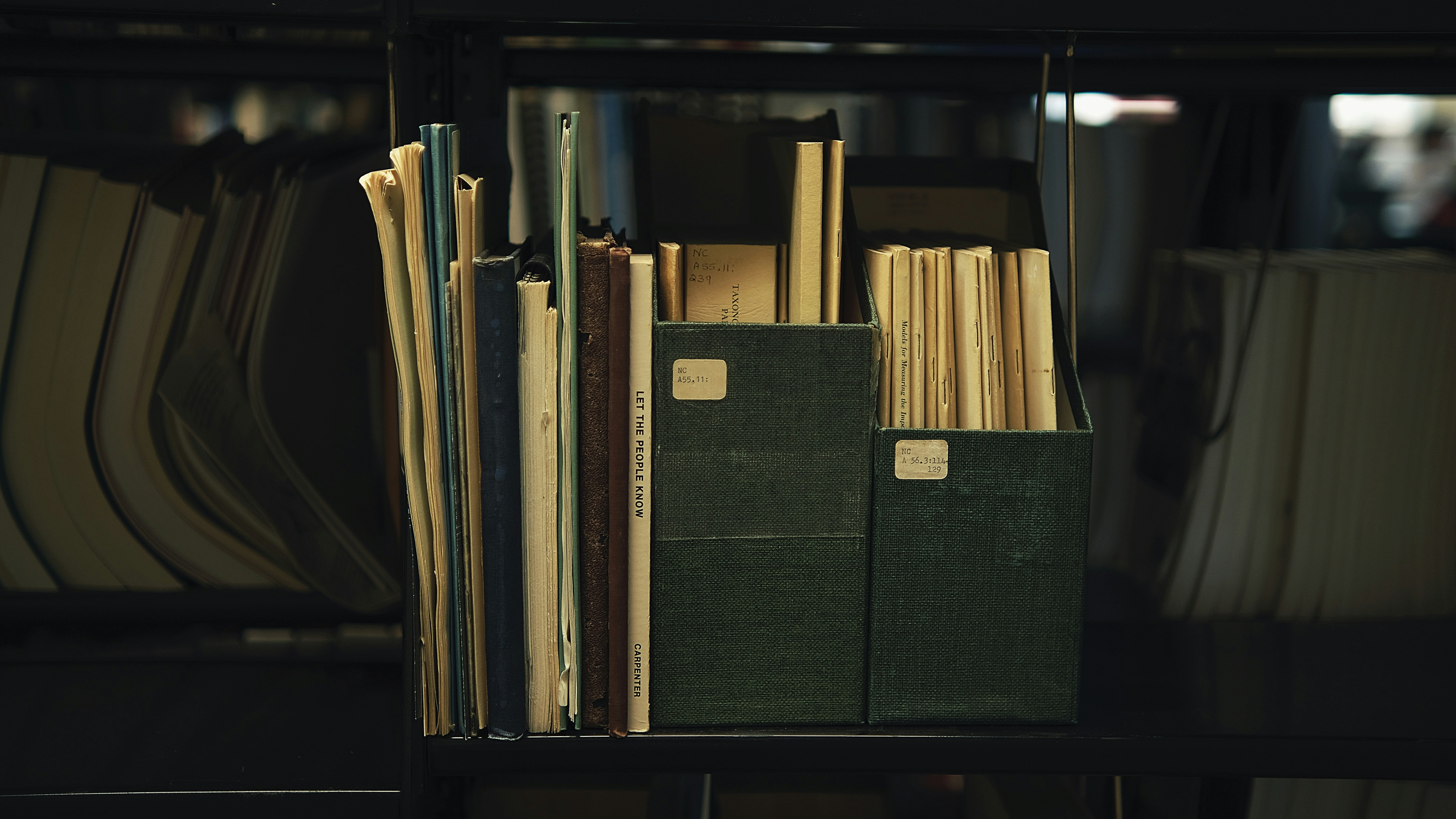 A dark archival shelf of aged books, folders, and labeled storage boxes lit in muted low light