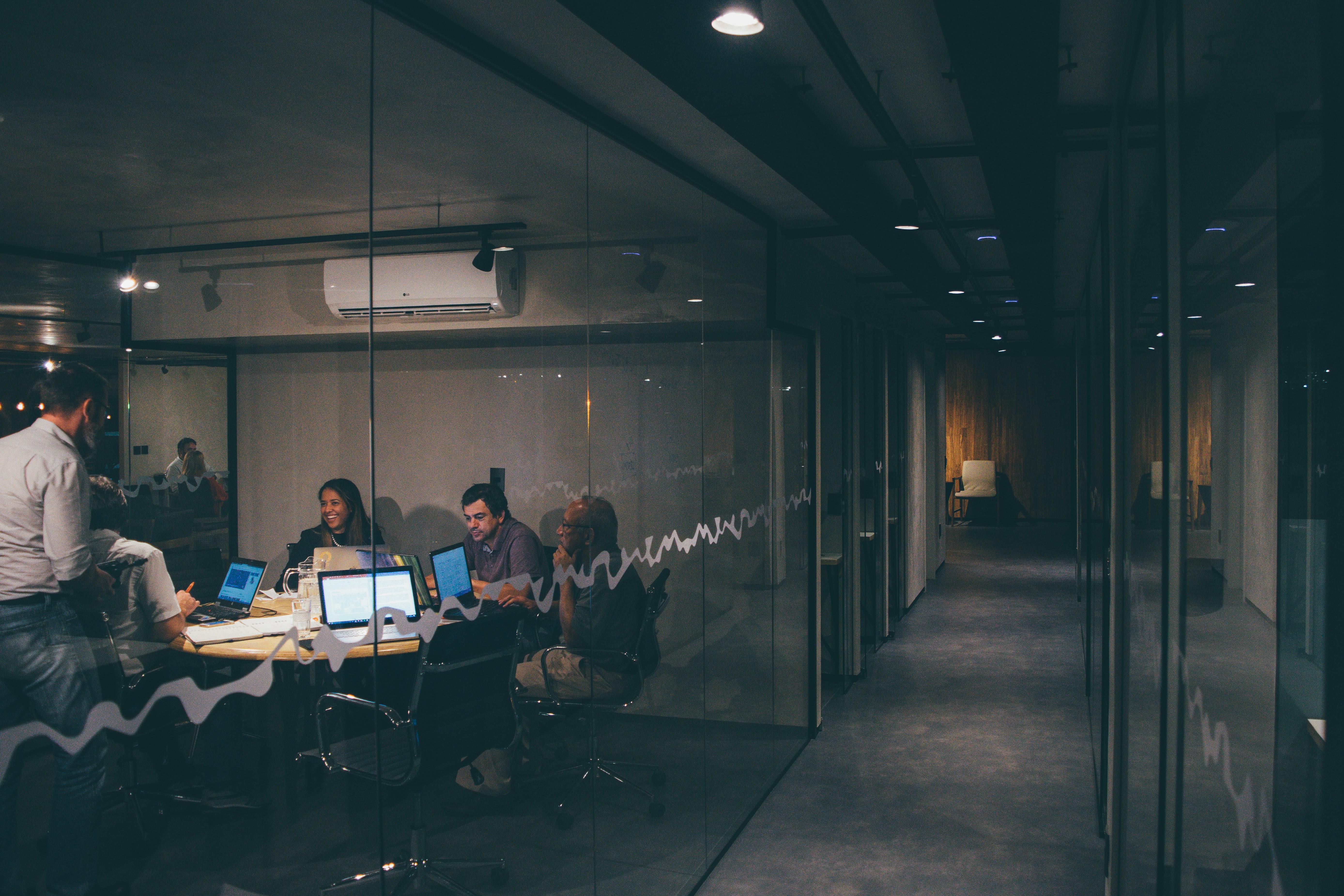 A team working late around a table in a glass-walled meeting room, viewed from an empty corridor