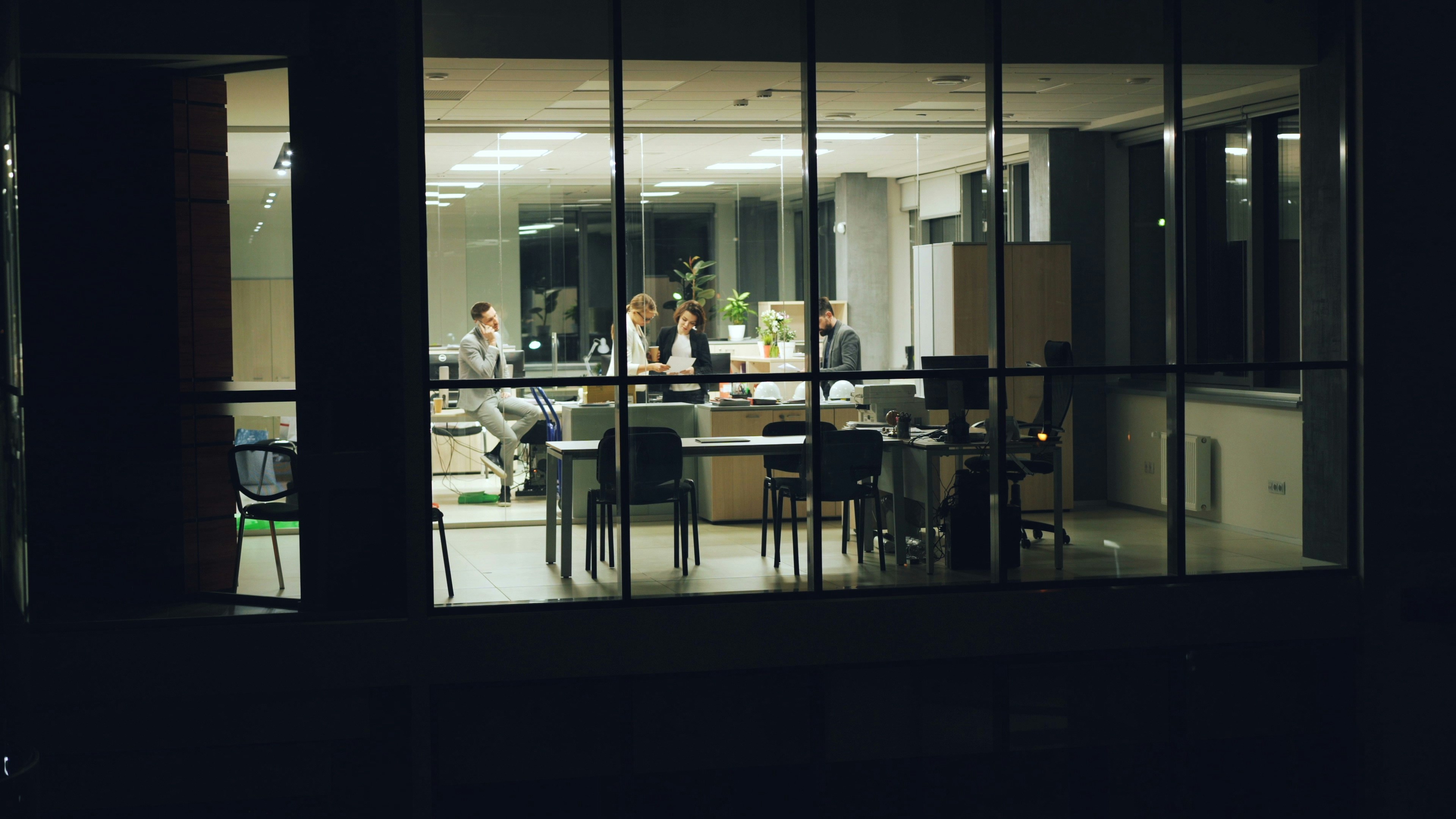 A lit modern open-plan office seen at night through a dark glass wall, with colleagues working late at their desks