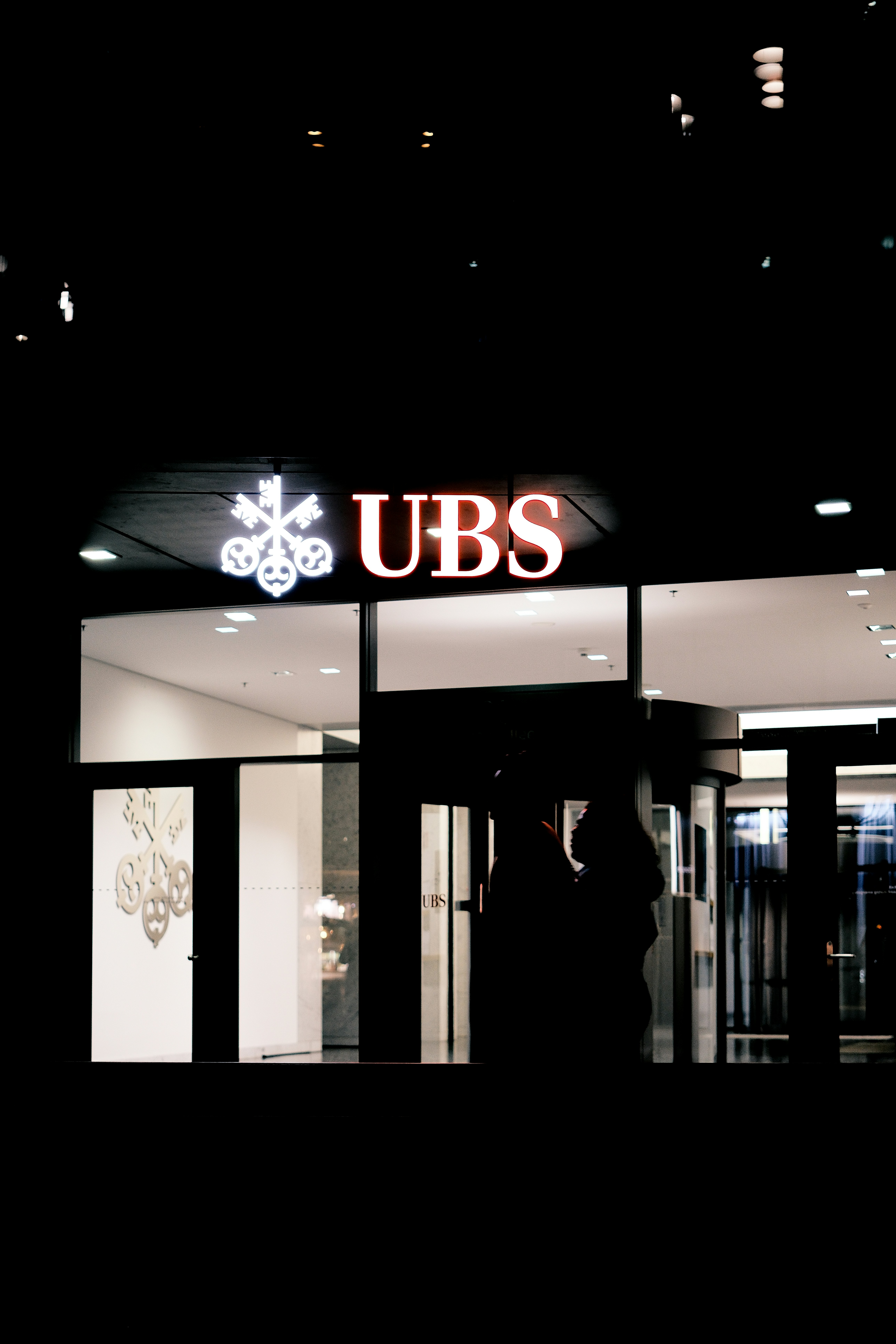 The illuminated red UBS logo above a bank branch entrance at night, with a silhouetted figure near the doorway